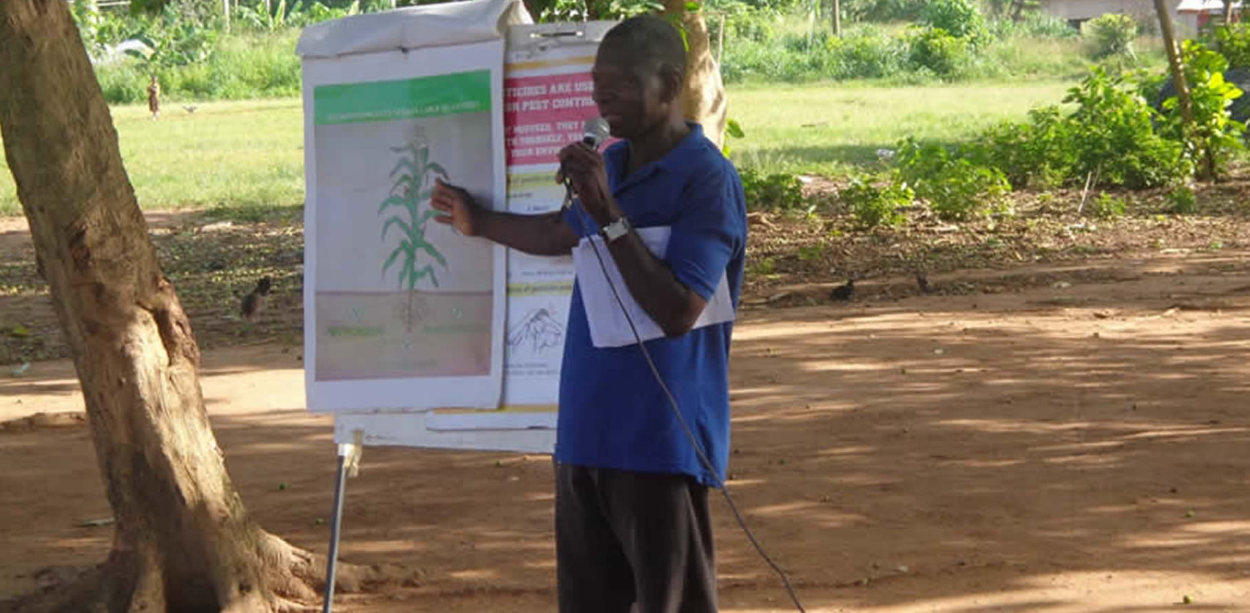 Community facilitator speaking into a microphone beside a flip chart outdoors in Ghana
